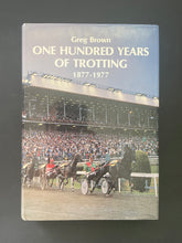 Load image into Gallery viewer, One Hundred Years of Trotting 1877-1977 by Greg Brown: photo of the front cover which shows minor scuff marks along the edges of the dust jacket.
